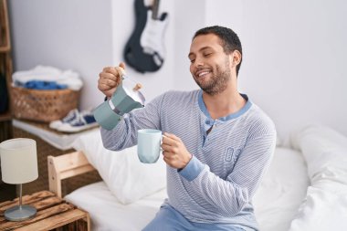 Young man drinking cup of coffee sitting on bed at bedroom