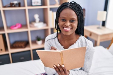 African american woman reading book sitting on bed at bedroom