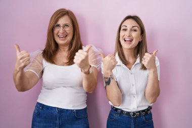 İspanyol anne ve kızı pembe arka plan işareti üzerine günlük beyaz t-shirt giyiyorlar. Elleriyle olumlu jest yapıyorlar. Gülümsüyorlar ve mutlular. Neşeli ifade ve kazanan jest. 
