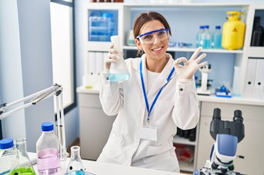 Young brunette woman working at scientist laboratory doing ok sign with fingers, smiling friendly gesturing excellent symbol 