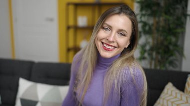 Sarışın genç bir kadın evdeki koltukta kendinden emin bir şekilde oturarak gülümsüyor.