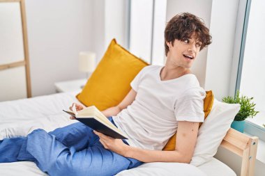 Young hispanic man reading book sitting on bed at bedroom