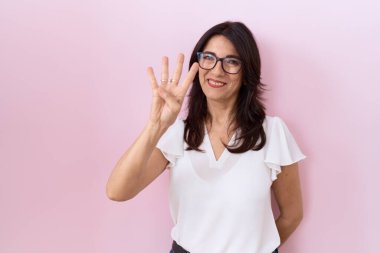 Orta yaşlı İspanyol kadın, günlük beyaz tişört ve gözlük giyip dört numaralı parmaklarıyla işaret ederken kendinden emin ve mutlu bir şekilde gülümsüyor.. 