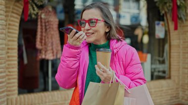 Gri saçlı, gülümseyen, alışverişe giden, giyim mağazasında akıllı telefonuyla sesli mesaj gönderen olgun İspanyol kadın.