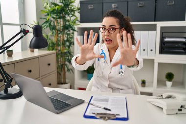 Young hispanic woman wearing doctor uniform and stethoscope afraid and terrified with fear expression stop gesture with hands, shouting in shock. panic concept. 