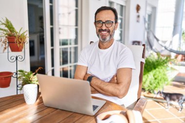 Orta yaşlı bir adam evde bilgisayar kullanıyor. Kollarını kavuşturup kameraya bakarken gülümsüyor. pozitif kişi. 