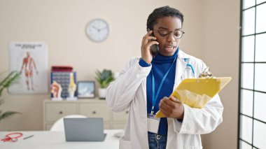 Afro-Amerikalı kadın doktor. Panoda belge okuyor. Klinikte akıllı telefondan konuşuyor.