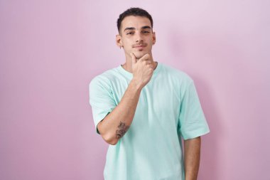 Pembe arka planda duran yakışıklı İspanyol adam kameraya bakarken kollarını kavuşturup, ellerini çenesine kaldırarak gülümsüyor. Olumlu düşünmek. 