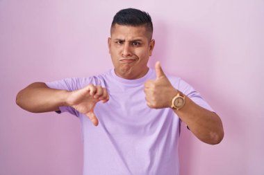 Young hispanic man standing over pink background doing thumbs up and down, disagreement and agreement expression. crazy conflict 