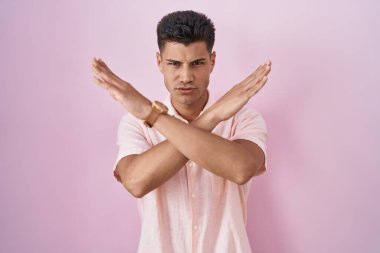 Genç İspanyol bir adam pembe arka planda duruyor. Reddedilme ifadesi, kollarını kavuşturma, negatif işaret, kızgın surat. 