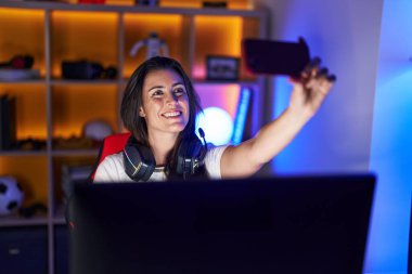 Young beautiful hispanic woman streamer smiling confident make selfie by smartphone at gaming room