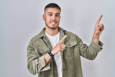 Genç İspanyol adam izole edilmiş arka planda duruyor gülümsüyor ve kameraya bakıyor iki eliyle ve parmaklarıyla kenarı işaret ediyor.. 