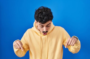 Mavi arka planda dikilen İspanyol adam parmaklarını göstererek reklamını yapıyor, şaşırmış yüz ve ağzını açıyor. 