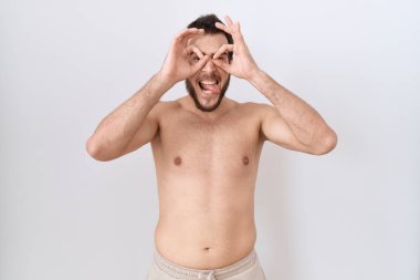 Beyaz arka planda üstsüz duran İspanyol genç adam dürbün gibi jest yapıyor, gözleri parmaklarından bakıyor. çılgın ifade. 