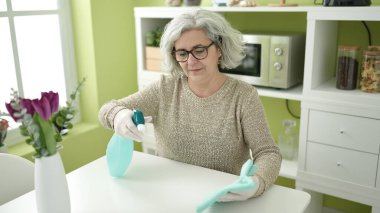 Middle age woman with grey hair cleaning table at home