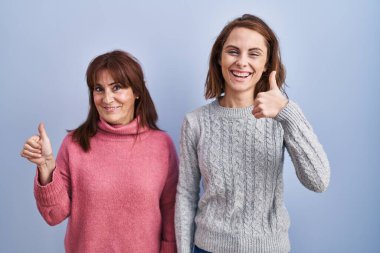 Anne ve kızı mavi arka planda durup mutlu bir el hareketi yapıyorlar. Kameraya bakıp başarıyı gösteren ifade. 