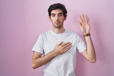 Young hispanic man standing over pink background swearing with hand on chest and open palm, making a loyalty promise oath 