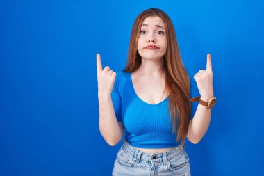 Mavi arka planda dikilen kızıl saçlı kadın üzgün ve üzgün görünüyor, parmaklarıyla yön gösteriyor, mutsuz ve depresif.. 
