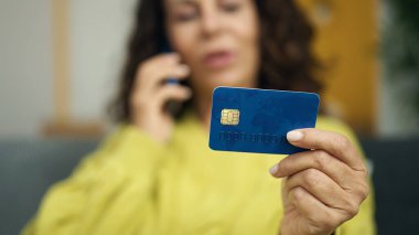 Middle age hispanic woman talking on smartphone holding credit card at home