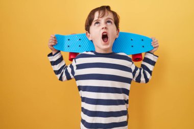 Küçük beyaz çocuk sarı arka planda kaykay tutuyor kızgın ve kızgın çığlık atıyor sinirli ve öfkeli, öfkeyle bağırıyor.. 