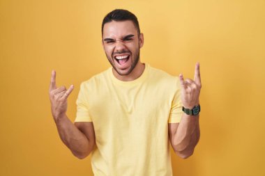 Young hispanic man standing over yellow background shouting with crazy expression doing rock symbol with hands up. music star. heavy music concept. 