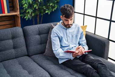 Evdeki kanepede oturan Latin kökenli genç adam deftere yazı yazıyor.