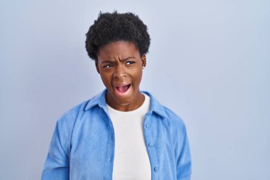 African american woman standing over blue background angry and mad screaming frustrated and furious, shouting with anger. rage and aggressive concept. 