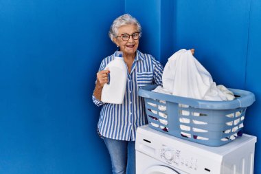 Çamaşır odasında elinde deterjan şişesiyle gülümseyen yaşlı gri saçlı kadın.