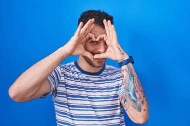 Mavi arka planda duran, elleri ve parmaklarıyla kalp şeklinde gülümseyen İspanyol genç adam tabelanın içinden bakıyor. 