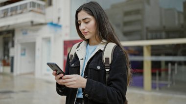 Genç, güzel, İspanyol bir kız öğrenci sokakta rahat bir ifadeyle akıllı telefon kullanıyor.