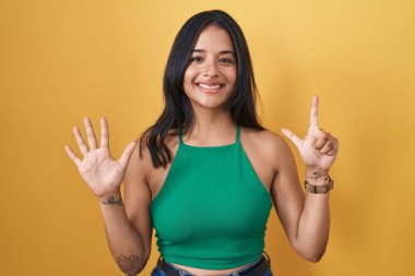 Sarı arka planda duran esmer kadın yedinci parmaklarını gösterip işaret ederken kendinden emin ve mutlu bir şekilde gülümsüyor.. 