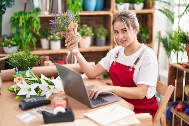 Young blonde woman florist using laptop holding lavender plant at flower shop