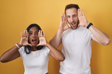 Interracial couple standing over yellow background smiling cheerful playing peek a boo with hands showing face. surprised and exited 