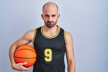 Basketbol forması giyen sakallı genç kel adam topa şüpheci ve gergin bakıyor, sorun yüzünden suratını asıyor. negatif kişi. 