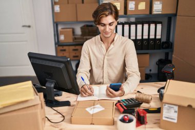 Young caucasian man ecommerce business worker using smartphone writing on notebook at office