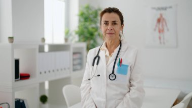 Orta yaşlı İspanyol kadın doktor üniforması giyiyor klinikte rahat bir ifadeyle dikiliyor.