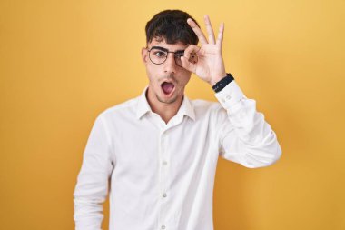 Young hispanic man standing over yellow background doing ok gesture shocked with surprised face, eye looking through fingers. unbelieving expression. 