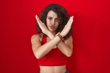 Kıvırcık saçlı İspanyol kadın kırmızı arka planda duruyor. Reddedilme ifadesi. Kollarını kavuşturup negatif işaret veriyor. 