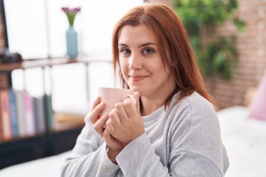Young beautiful plus size woman drinking cup of coffee sitting on bed at bedroom