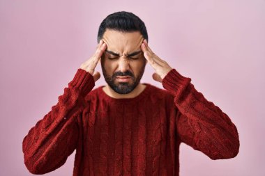 Sakallı genç İspanyol bir adam pembe arka planda rahat bir süveter giyiyor. Başı ağrıyor çünkü stres altında. migren ağrısı. 