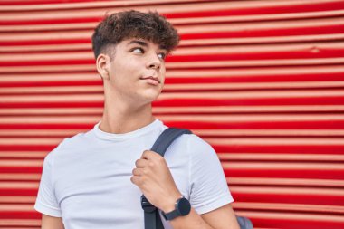 İzole edilmiş kırmızı metal arka planda gevşek bir ifadeyle sırt çantası takan İspanyol genç öğrenci.