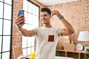 Young hispanic man having video call holding key of house at new home