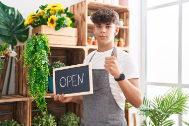 Çiçekçide çalışan İspanyol genç elinde açık tabela, mutlu ve olumlu gülümsüyor, başparmağıyla mükemmel ve onay işareti yapıyor. 