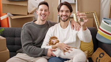 İki adam ellerinde anahtarlar, yeni evlerinde köpekleriyle kanepede oturuyorlar.
