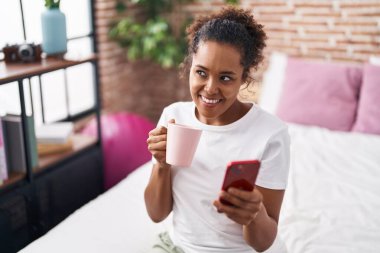 African american woman using smartphone drinking coffee at bedroom