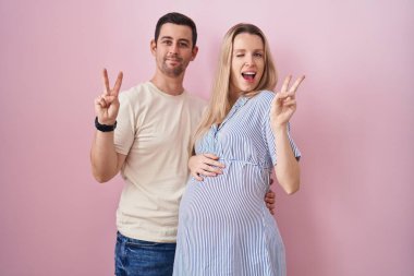 Genç bir çift pembe arka planda durup kameraya gülümseyen ve parmaklarıyla zafer işareti yapan bir bebek bekliyor. İki numara.. 