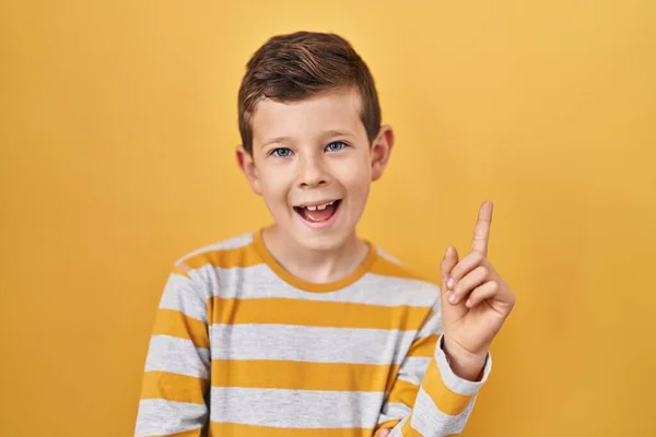 カメラを見ている側に手と指で指して 顔に大きな笑顔で黄色の背景の上に立って若い白人の子供 — ストック写真