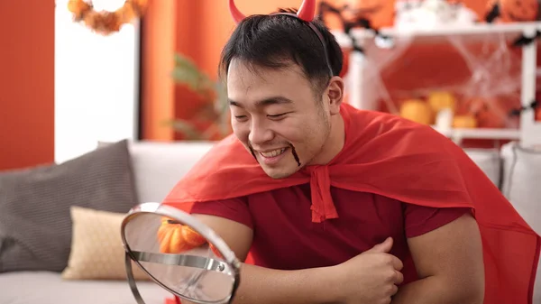 Young chinese man wearing devil costume looking on mirror at home