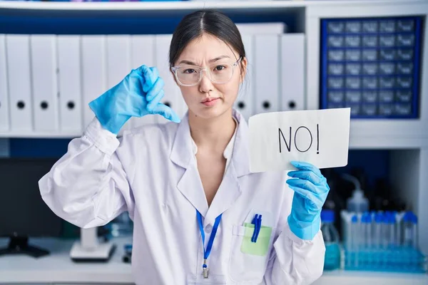 Chinese young woman working at scientist laboratory holding no banner with angry face, negative sign showing dislike with thumbs down, rejection concept