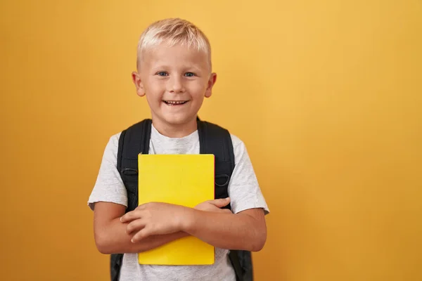 学生のバックパックを身に着けている小さな白人の少年と肯定的かつ幸せな立って見て本を保持し 歯を示す自信を持って笑顔で笑顔 — ストック写真